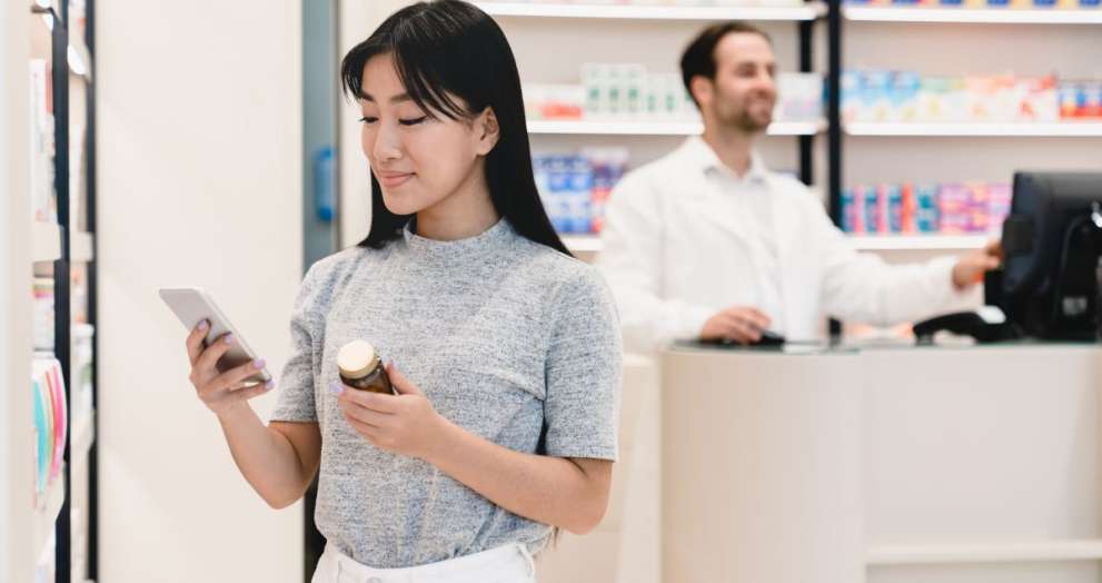 Female asian young client customer buyer checking information about side effects, ingredients, active substance in smart phone cellphone in drugstore pharmacy